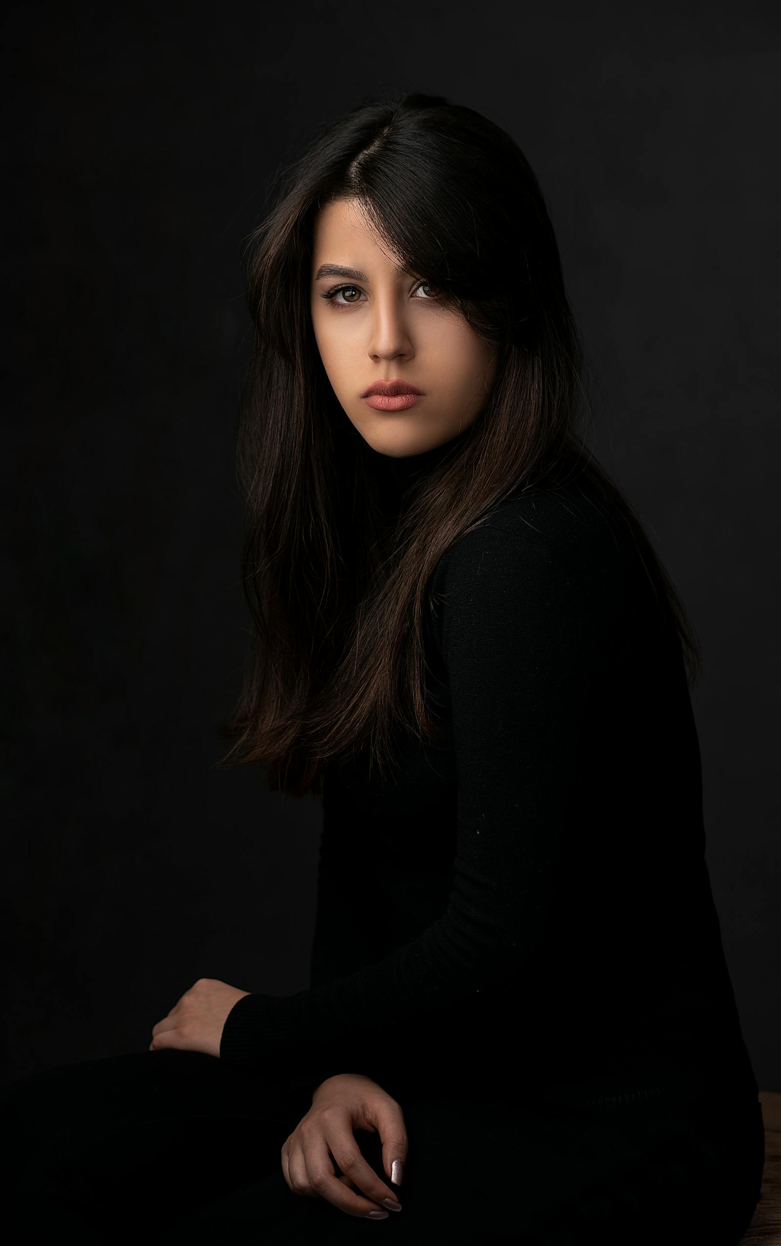 A studio portrait lit in warm tungsten — a young woman in profile against a deep charcoal seamless, soft fall-off on her shoulder, signature JAMES VISUALS lighting.